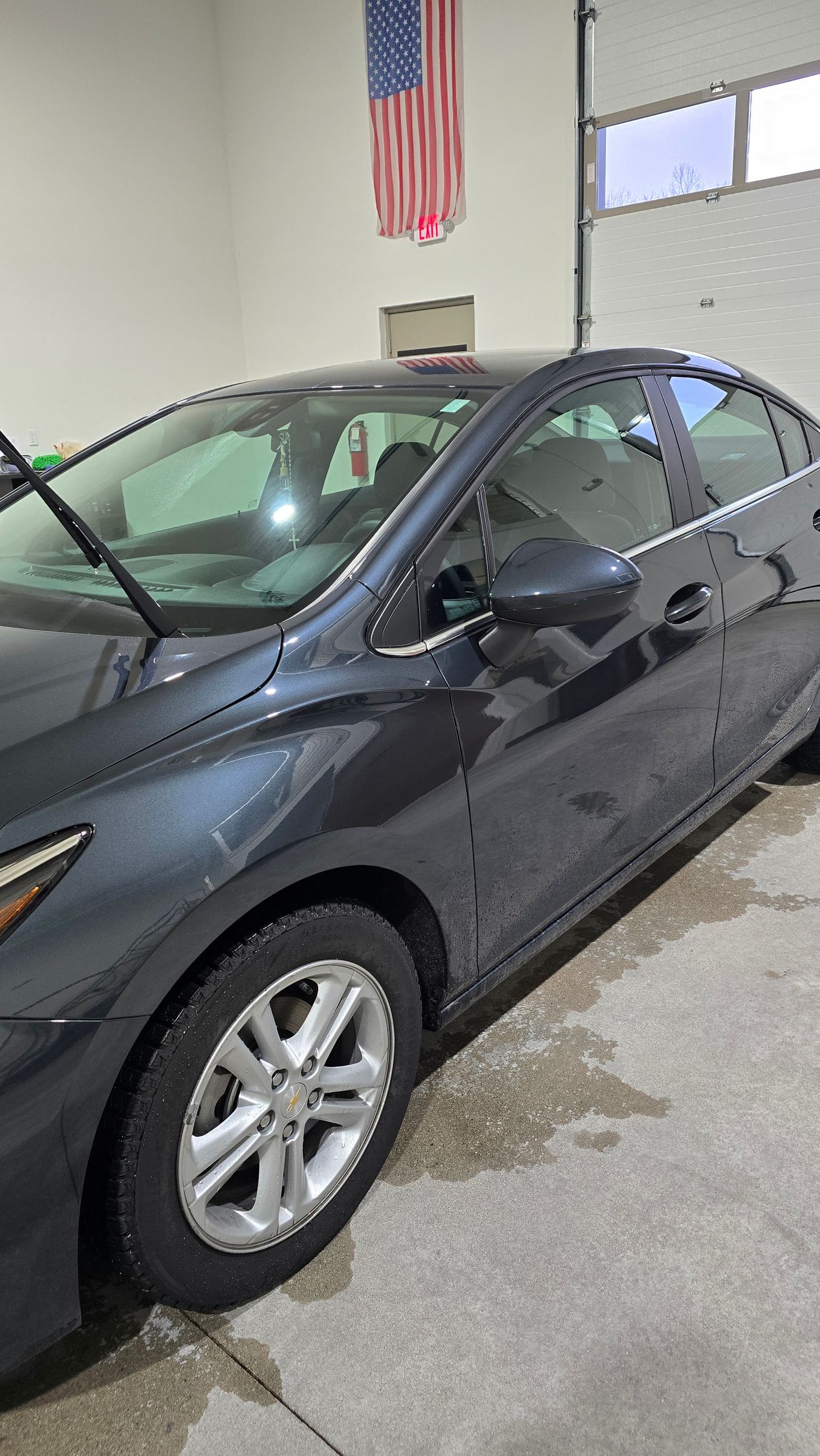 Dark gray sedan parked indoors in a garage or showroom, with an American flag on the wall.