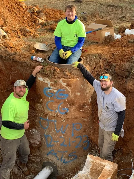Three workers in neon shirts pose around a giant excavated rock with blue markings at a dig site.