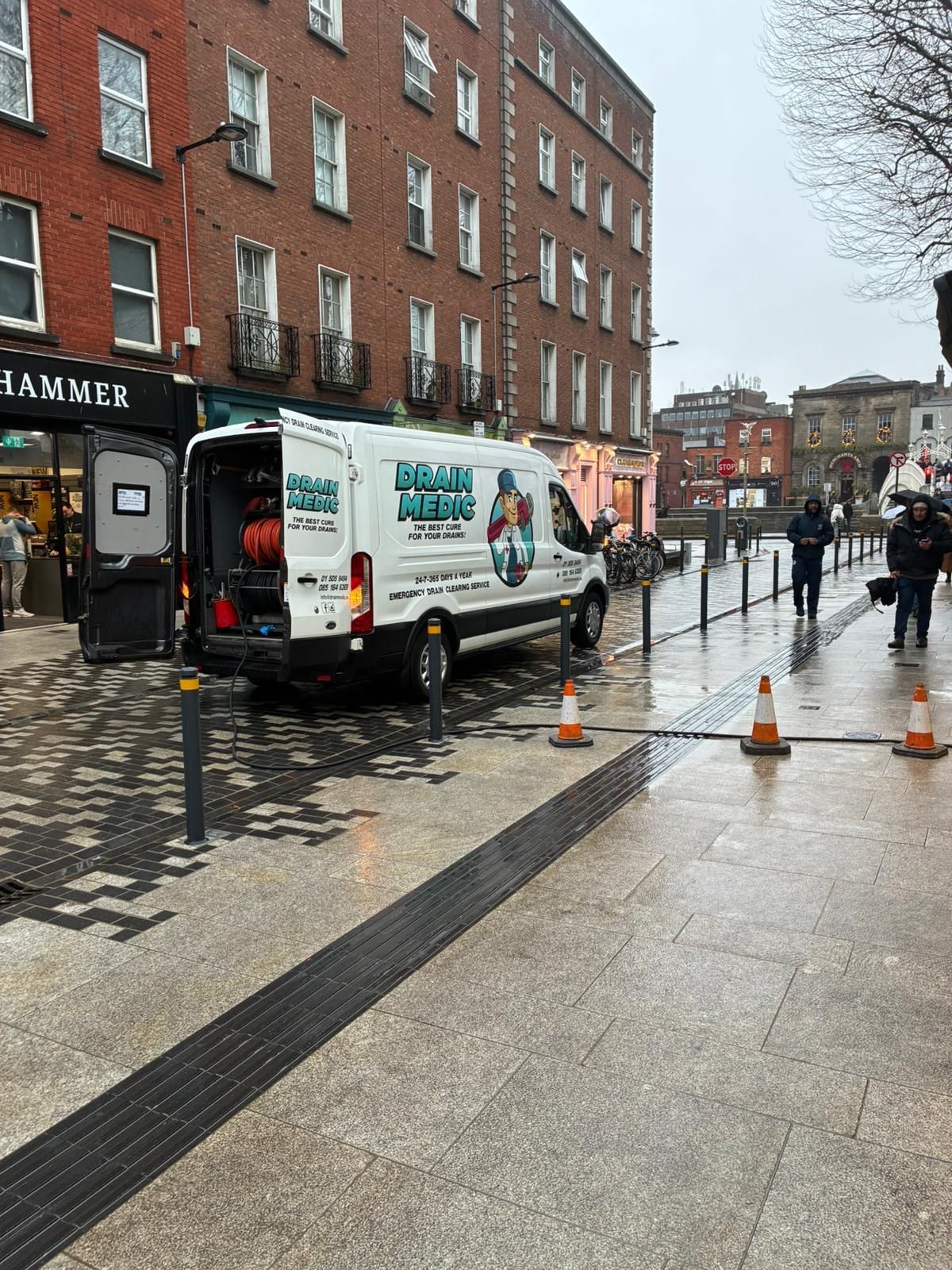 Van parked on wet city street beside brick buildings and orange cones, with a worker nearby