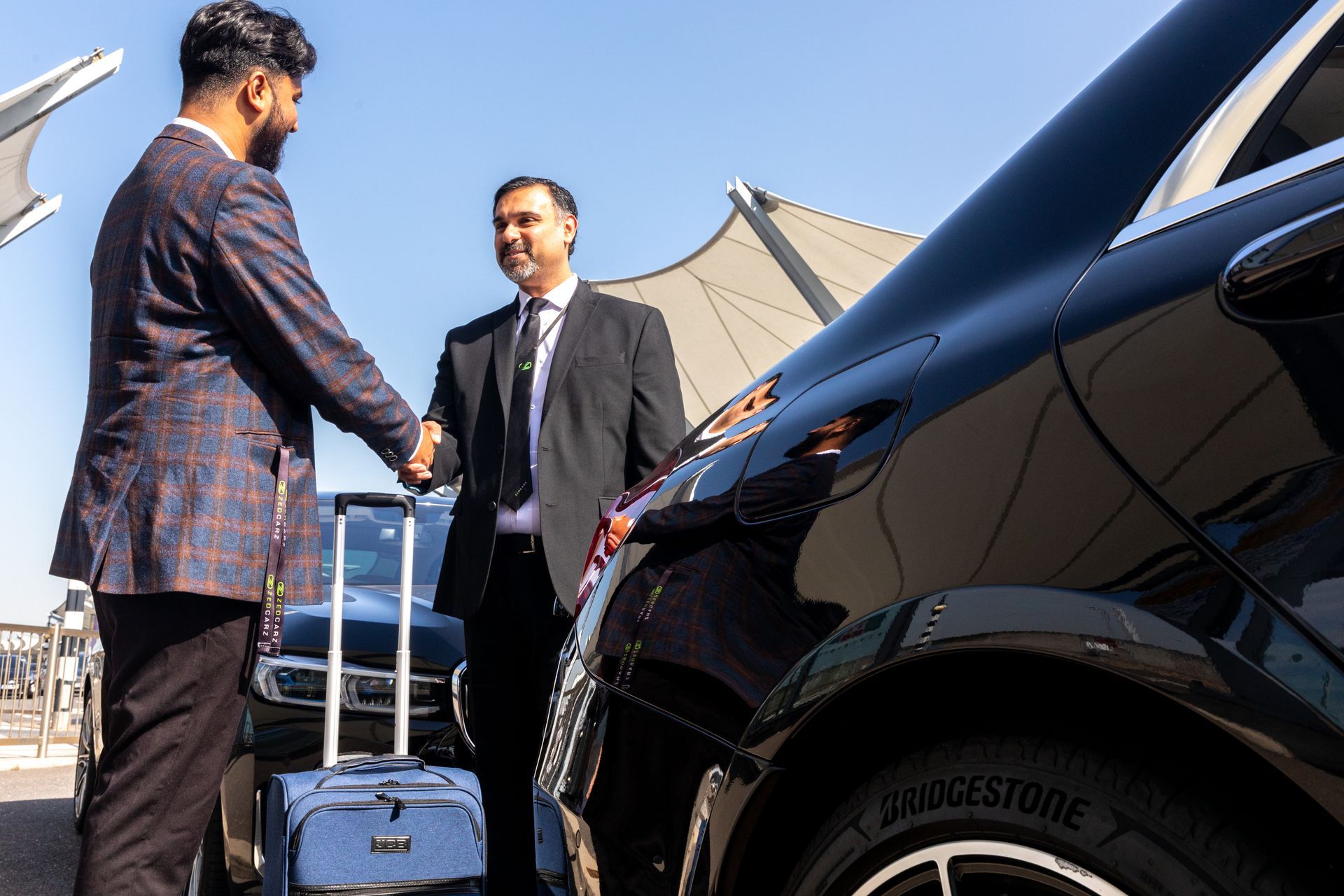 Two men shake hands beside a black car and luggage under a canopy.