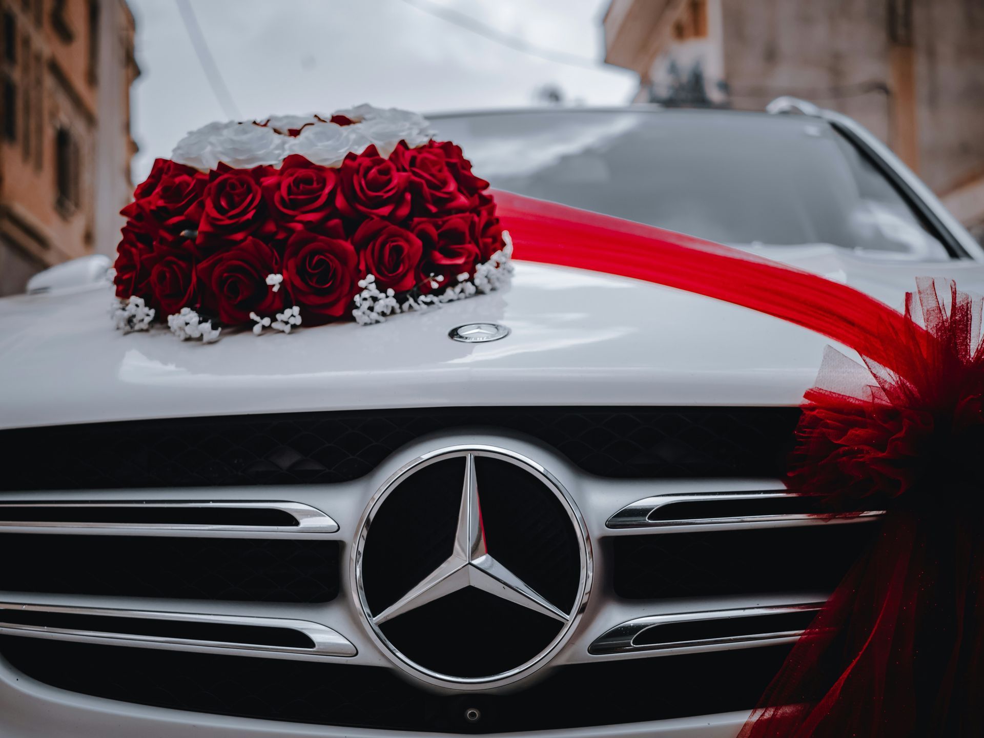 White Mercedes decorated with red roses and ribbon on the hood, parked on a city street