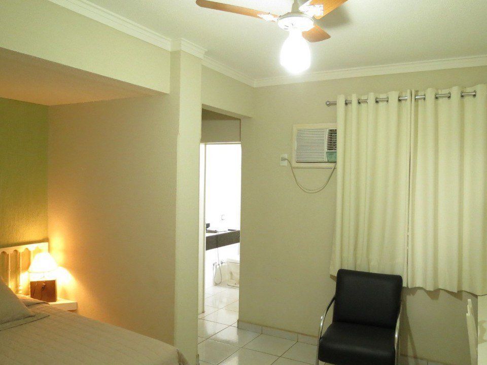Um quarto com uma cama, um abajur, uma cadeira preta, cortinas bege e uma porta que dá para um cômodo claro e branco.