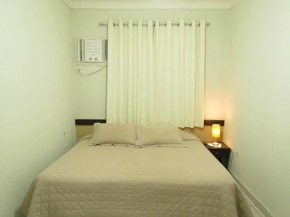 Um quarto simples, em tons neutros, com uma cama de casal arrumada, cortinas brancas, ar-condicionado de parede e um pequeno abajur.