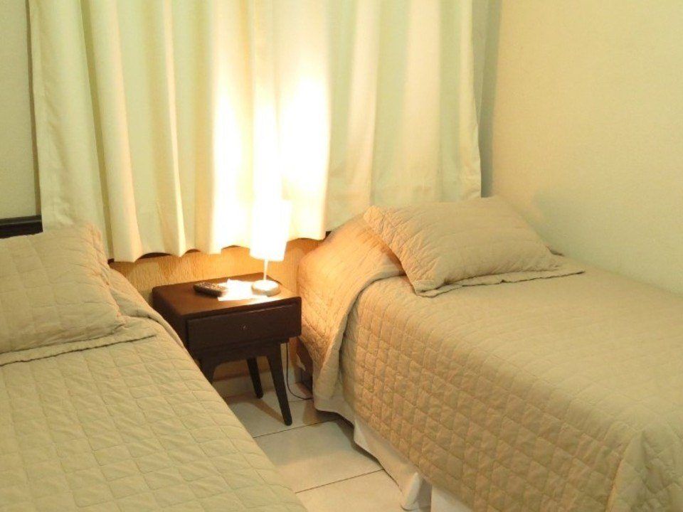 Um quarto de hotel com duas camas de solteiro, roupa de cama acolchoada em tons neutros de bege e uma pequena mesa de cabeceira com um abajur entre elas.