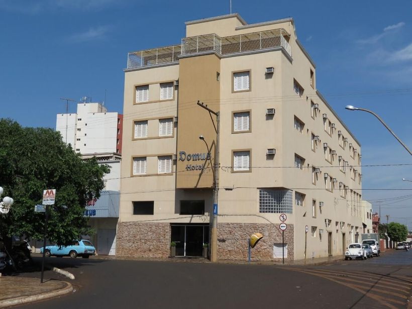 Um hotel bege de cinco andares numa esquina, com o térreo revestido de pedra e um céu azul ensolarado.