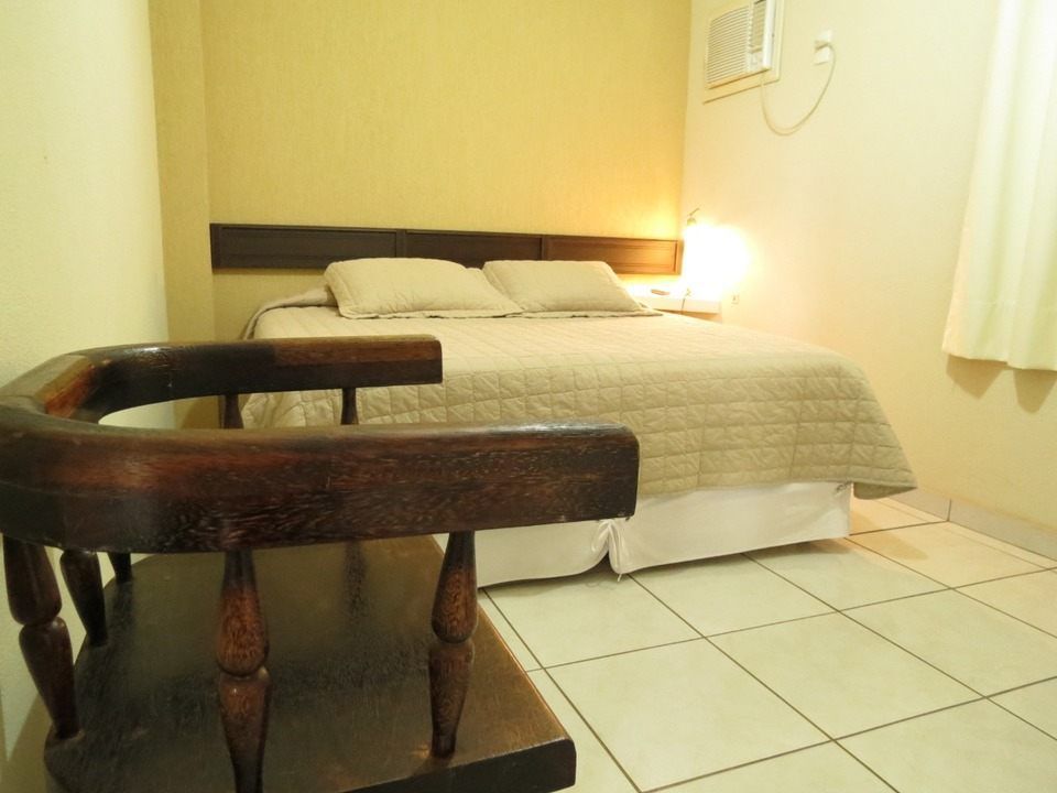 Uma cadeira de madeira está em primeiro plano em um quarto com cama arrumada, paredes claras e piso de azulejo.