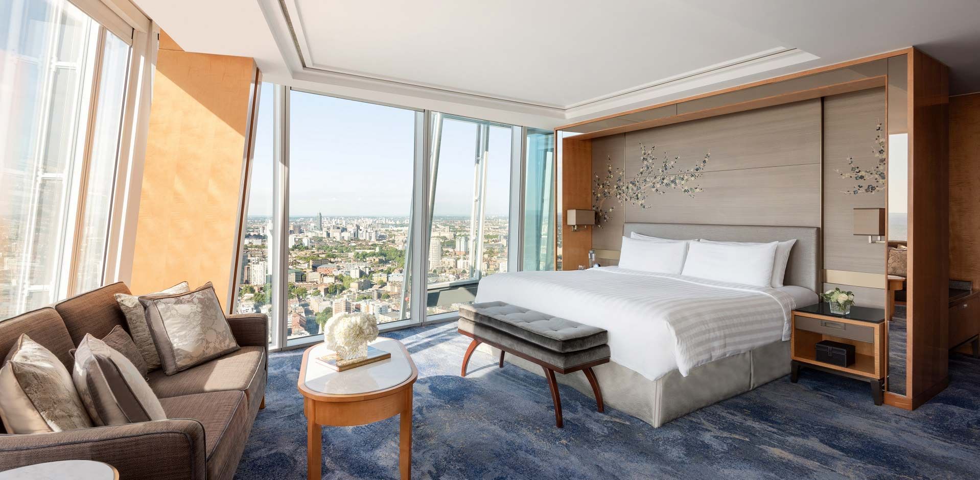 Um quarto de hotel moderno com uma cama grande, sofá marrom e janelas do chão ao teto que oferecem uma vista panorâmica da cidade.
