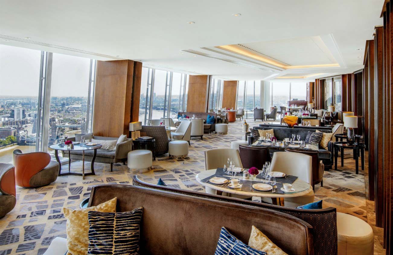 Interior elegante de restaurante com janelas do chão ao teto, vista para a cidade, assentos luxuosos e mesas postas para refeições.