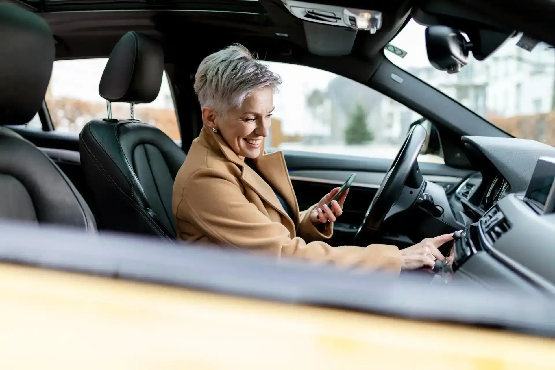 Donna in auto, sorridente, che usa il telefono e regola i comandi dell'auto. 