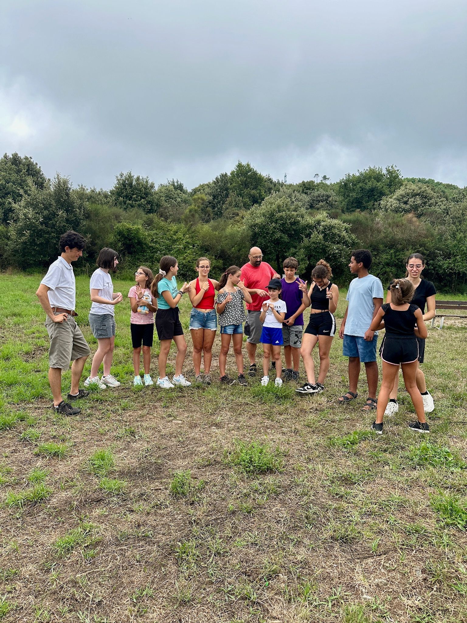 Gruppo di persone in piedi all'aperto, su un campo erboso, vicino agli alberi, sotto un cielo coperto.