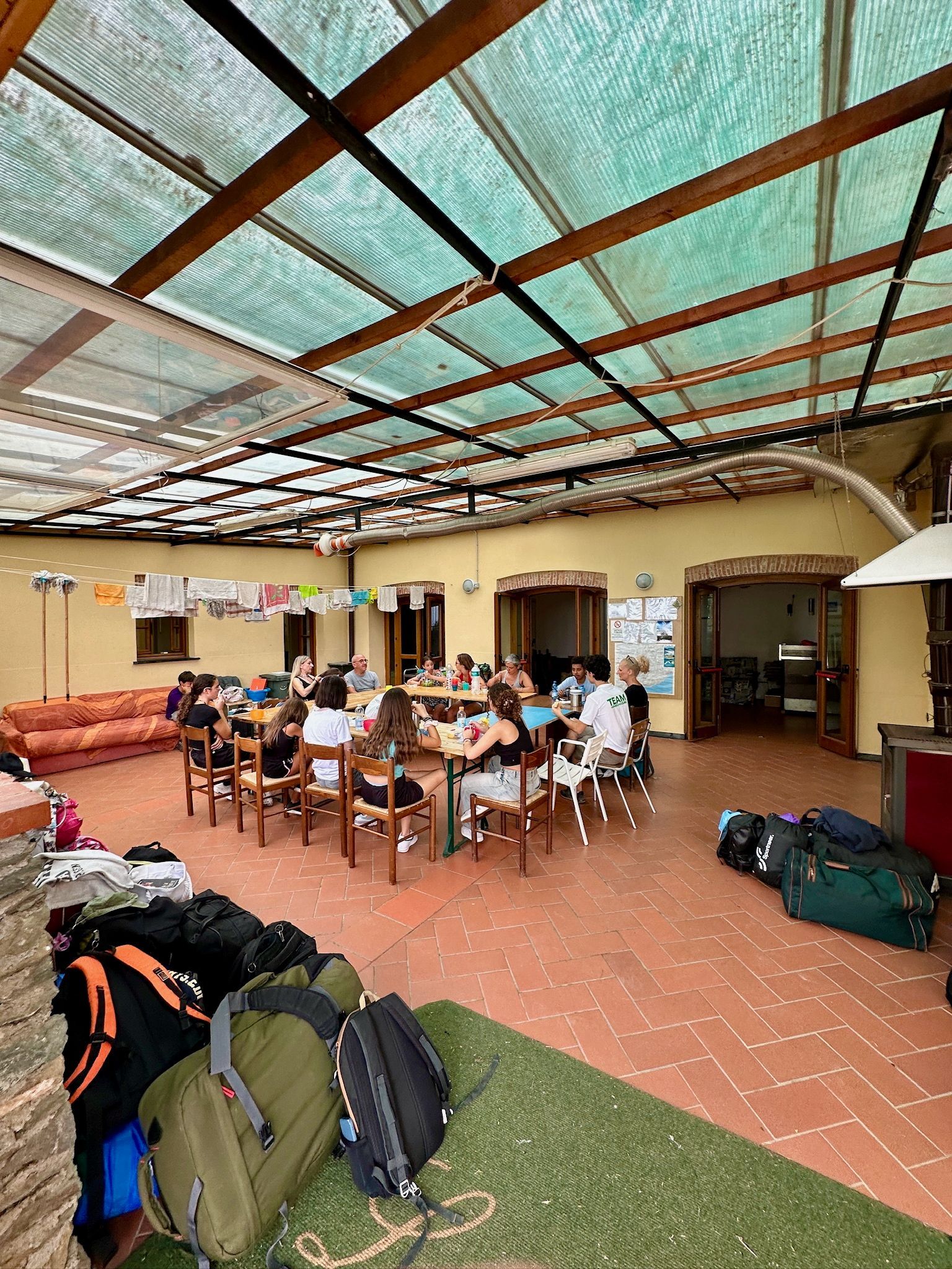Persone sedute attorno a un grande tavolo, mentre cenano. Borse e bagagli sono ammucchiati lì vicino, sotto un patio coperto.