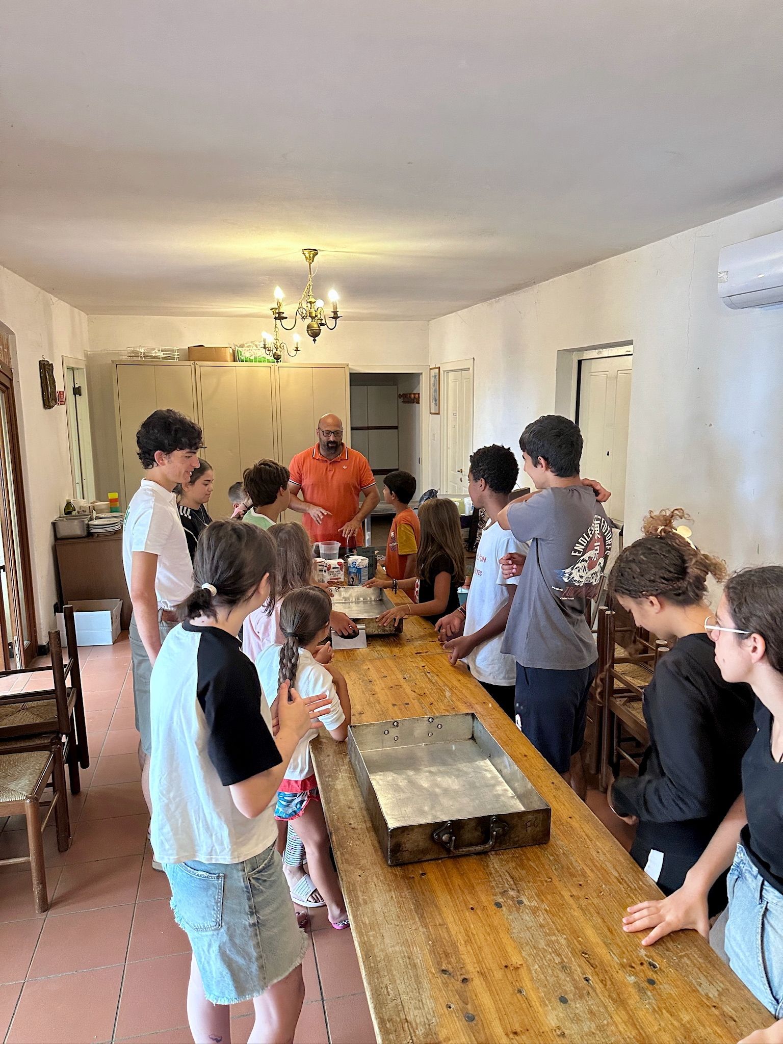 Un gruppo di persone attorno a un lungo tavolo di legno, con una persona che indossa una maglietta arancione. Al chiuso.