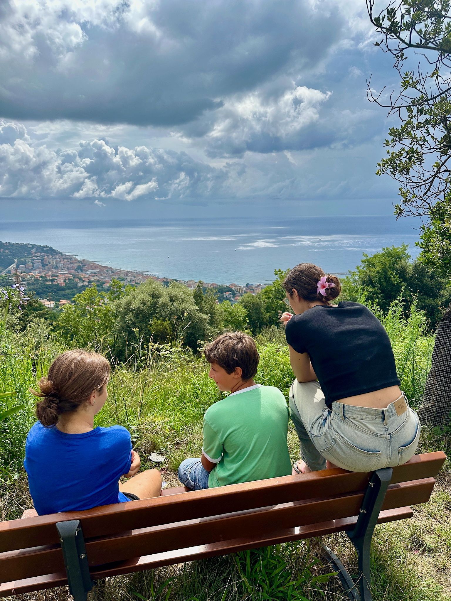 Tre persone sono sedute su una panchina con vista sulla città e sul mare. Il cielo è coperto.