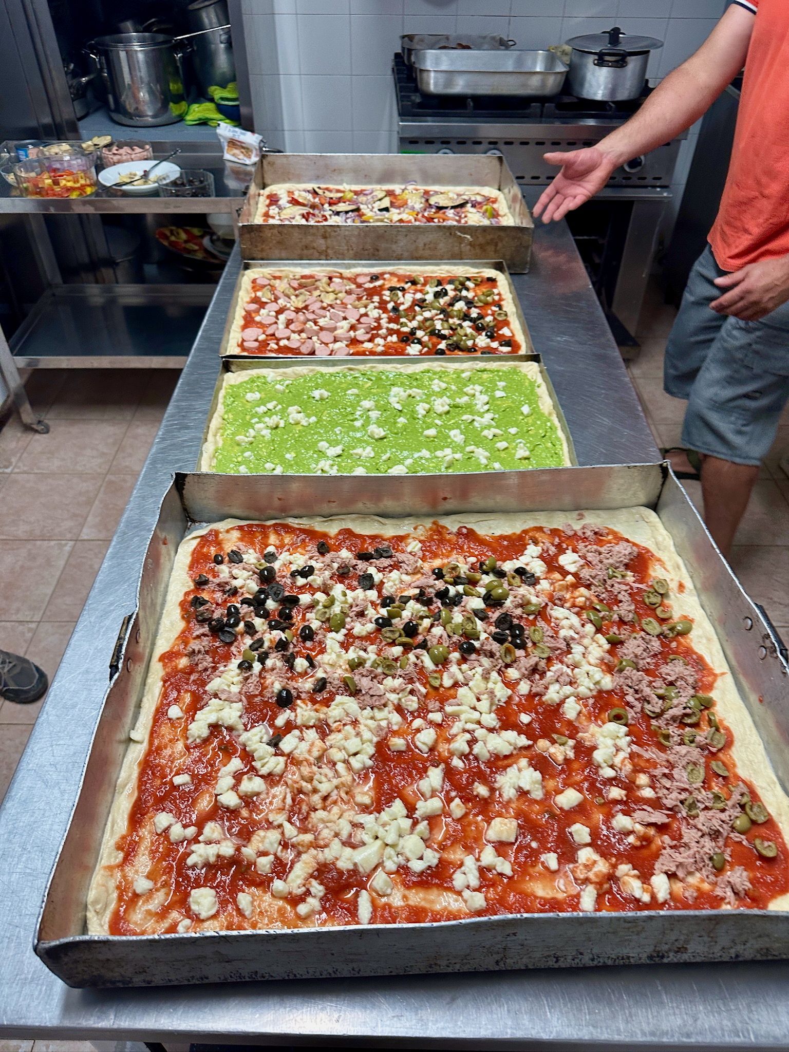 Quattro grandi pizze rettangolari in teglie di metallo sul bancone della cucina, con vari condimenti, una mano che ne indica una.