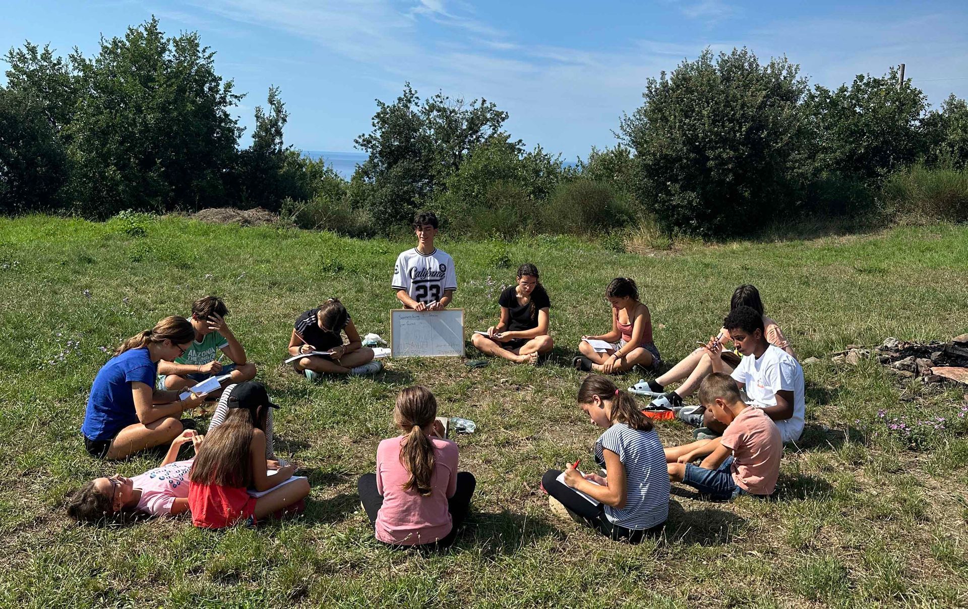 I bambini siedono in cerchio sull'erba, lavorando su dei fogli. Una persona è in piedi, con una lavagna bianca in mano. All'aperto, soleggiato.