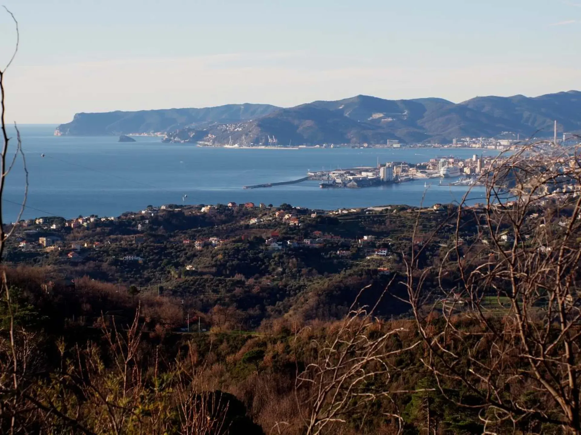 Vista costiera di una città sull'acqua con montagne sullo sfondo, alberi in primo piano e un cielo azzurro e limpido.