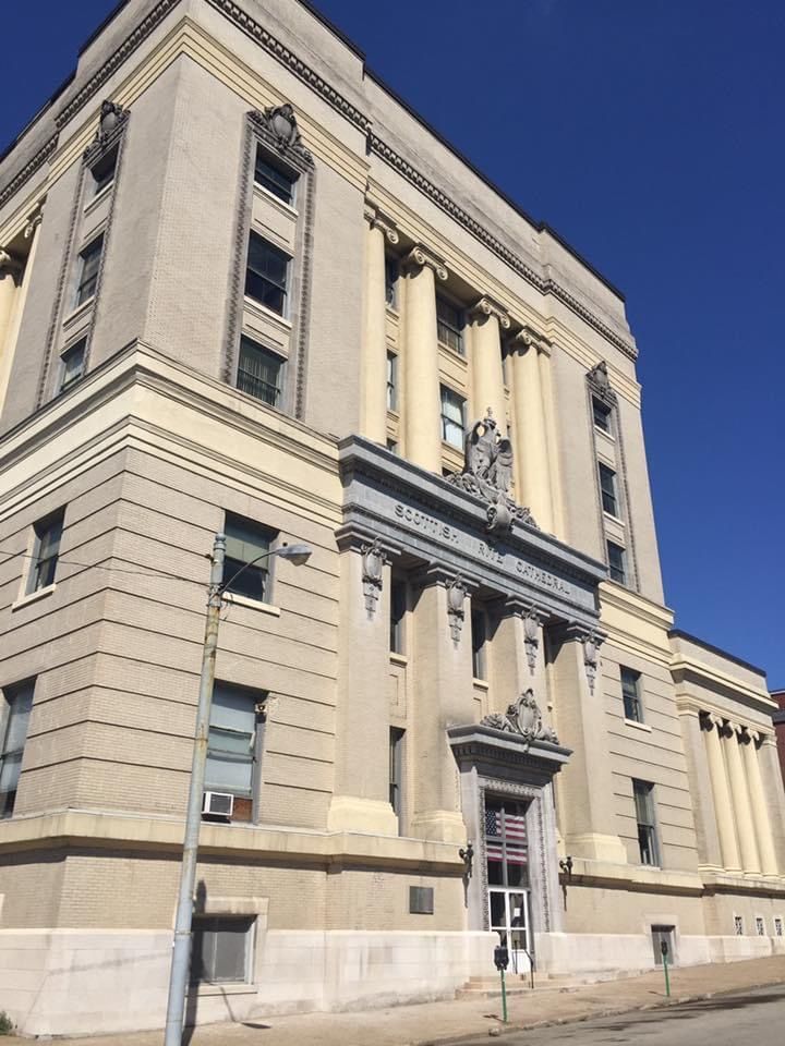 Exterior of a large, light-colored, neoclassical building with columns and a sign, set against a clear blue sky.