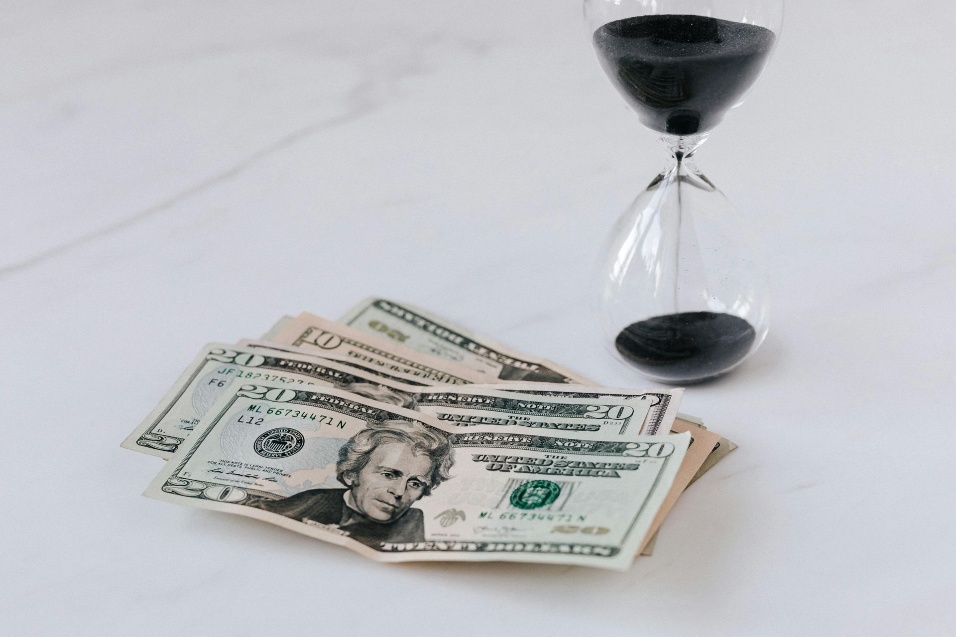 Pile of US dollar bills next to a nearly empty hourglass on a white surface.