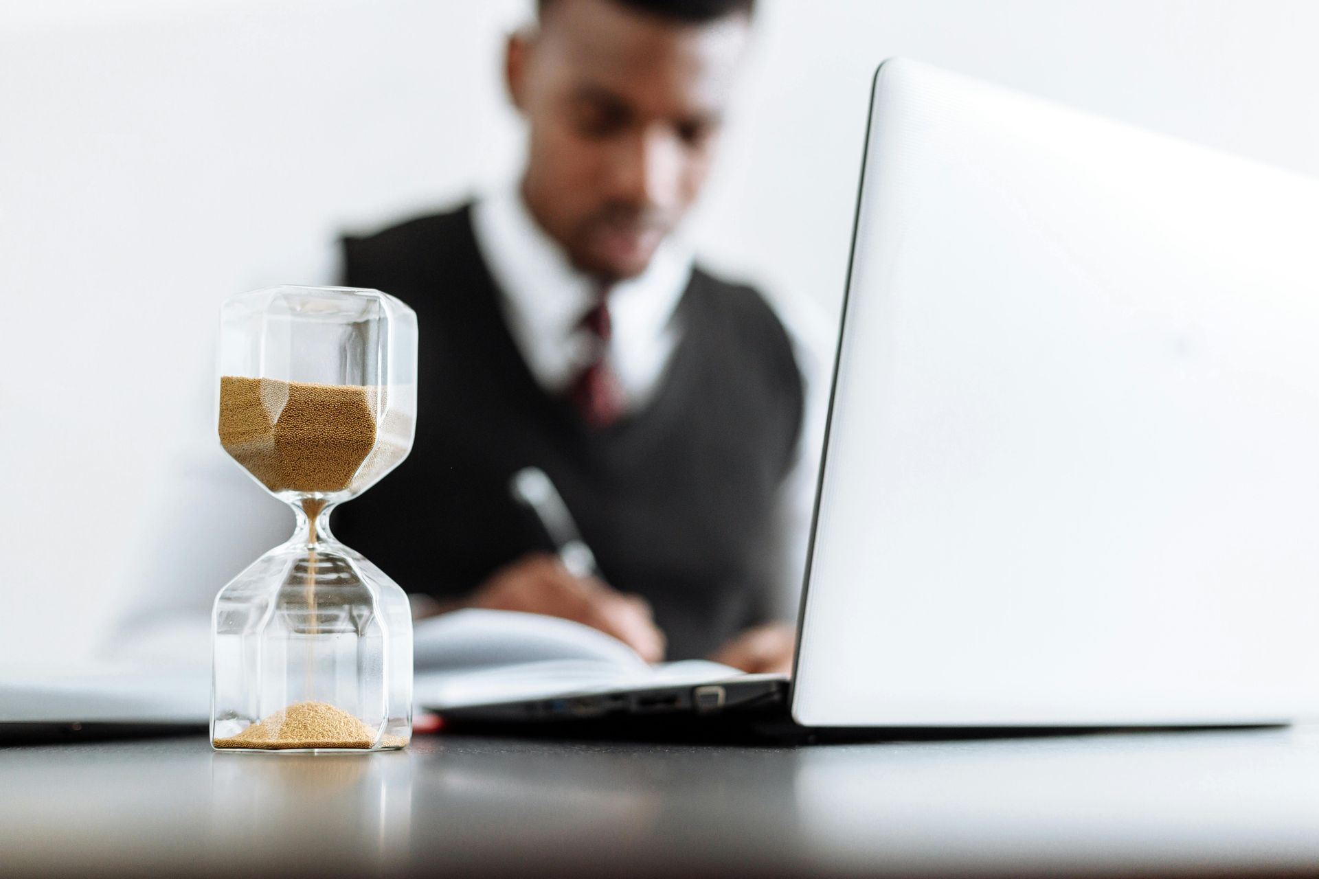 Hourglass on a desk with a laptop and a person in a suit working in the blurred background.