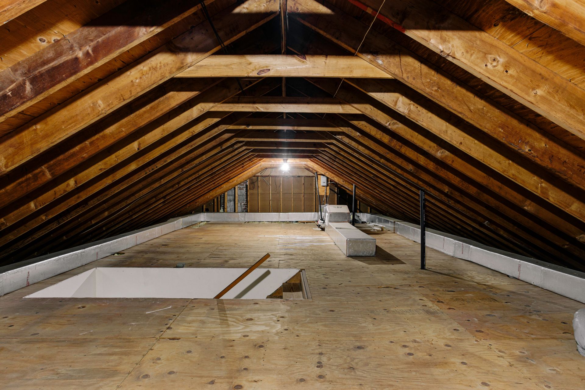 Unfinished attic with exposed wooden rafters, plywood floor, and scattered insulation panels