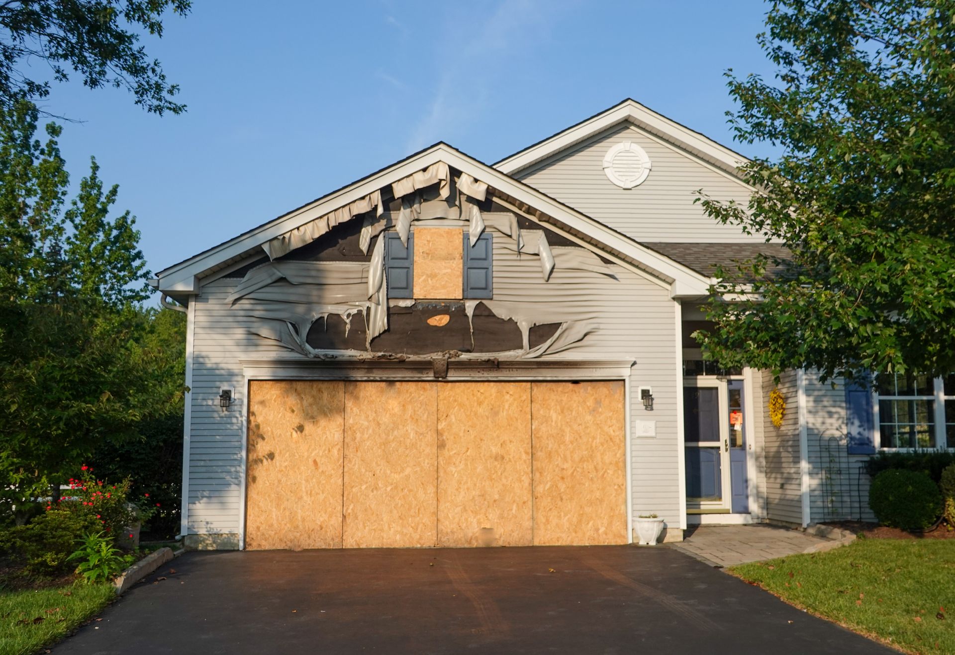 Boarded-up suburban house with damaged front porch and garage, viewed from the driveway.