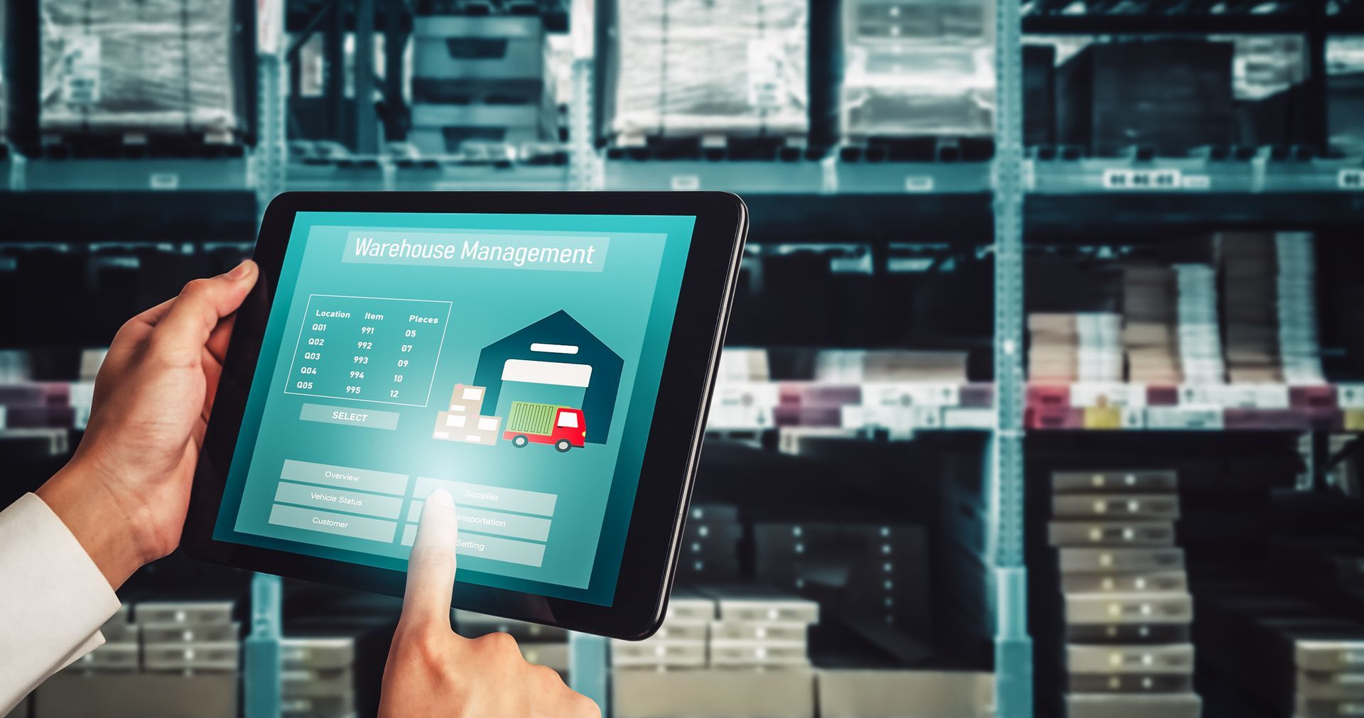 Hands using a tablet to monitor a warehouse inventory system with stacked boxes in the background
