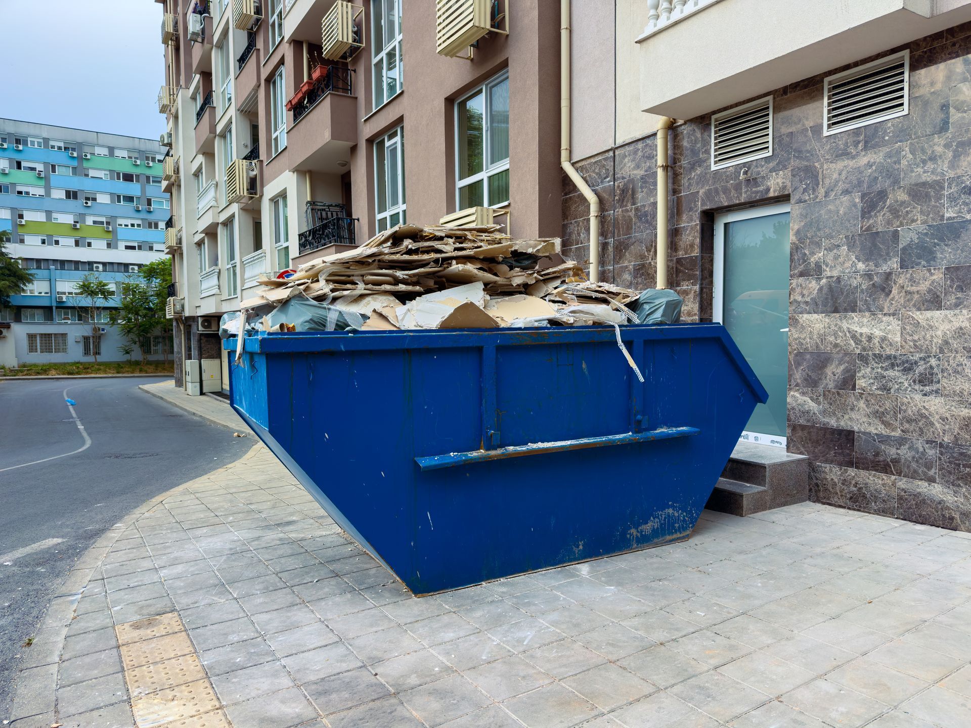 Large blue dumpster overflowing with debris on a sidewalk beside apartment buildings.