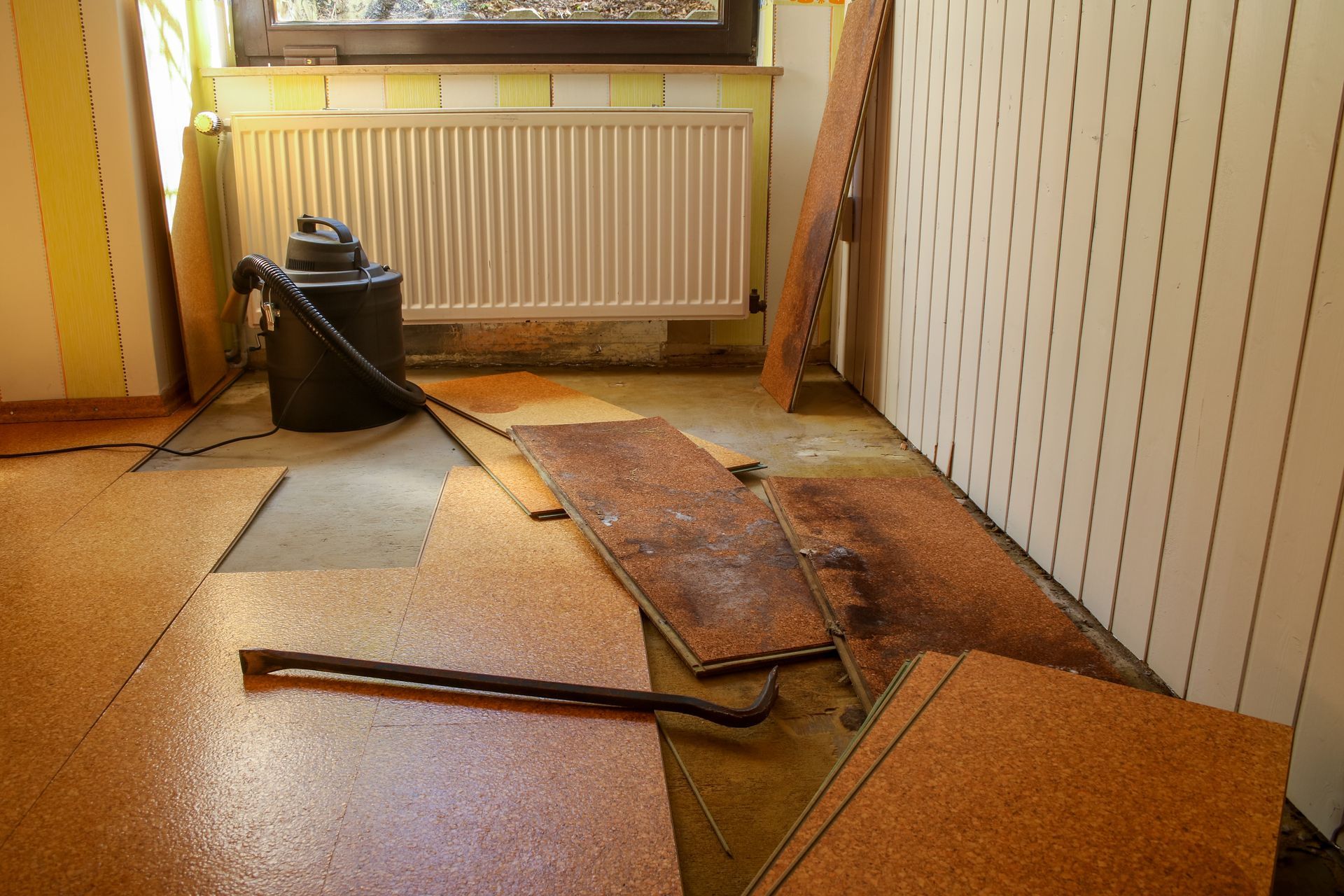 Damaged floor with broken boards and a canister vacuum in a small room near a radiator.