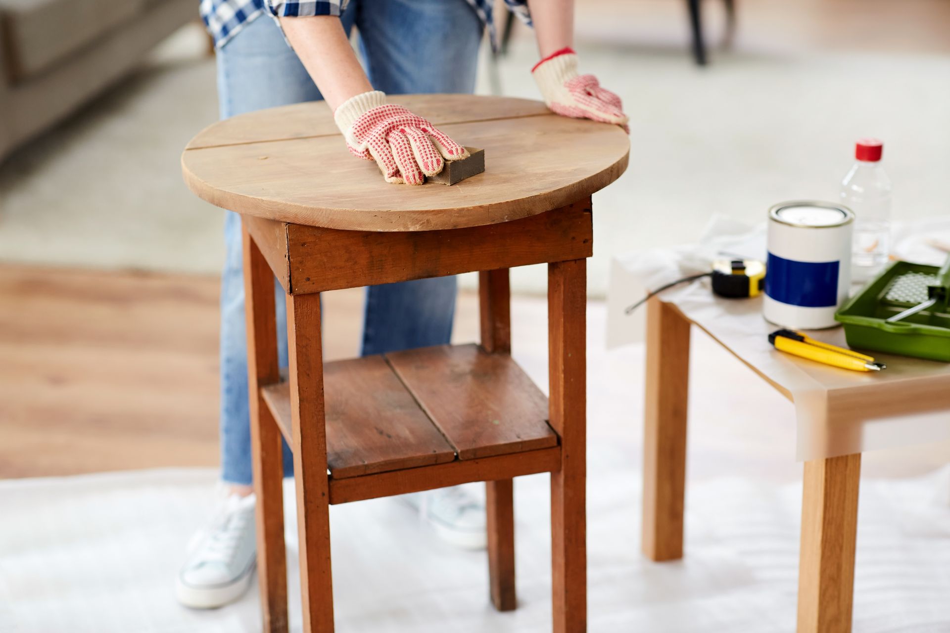 Person sanding a wooden table in a home workshop, with tools and paint supplies nearby