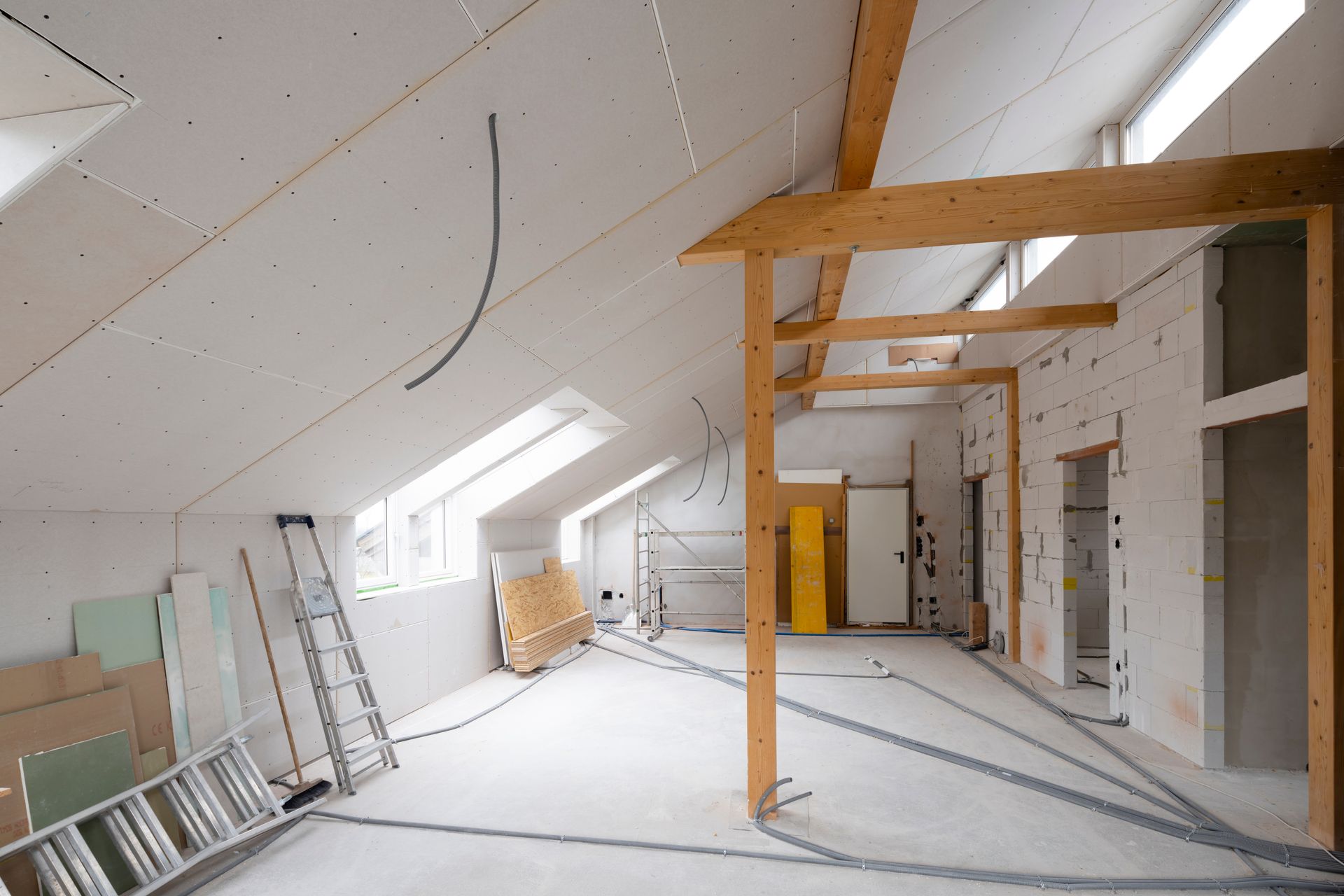 Empty attic under renovation with exposed wooden beams, ladders, and white slanted walls