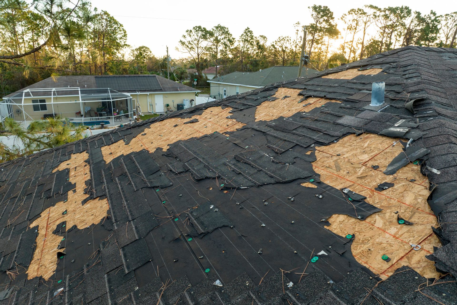 Damaged roof with missing shingles and exposed wood after storm, viewed at sunset.