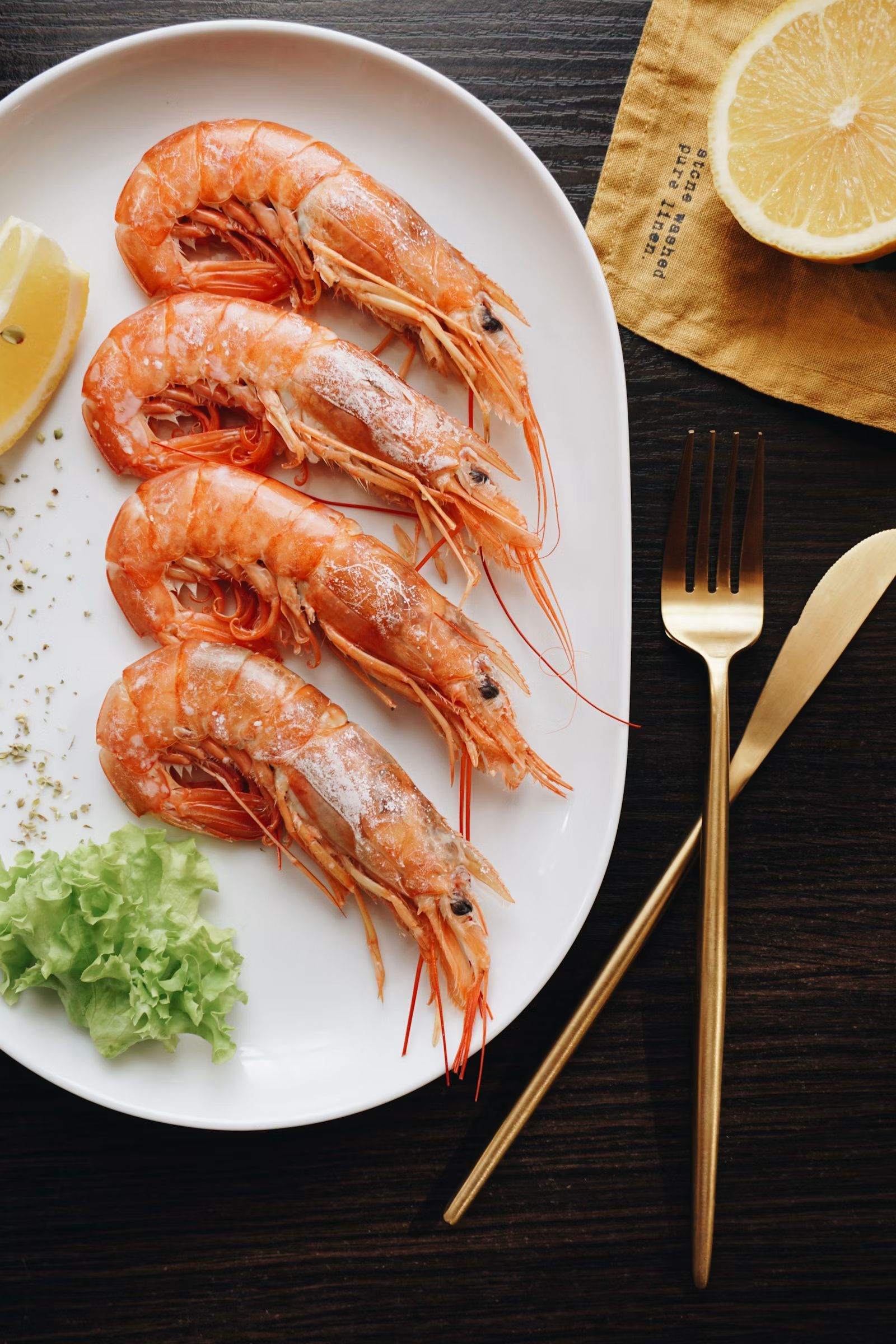 Plated cooked shrimp on a dark table with lemon, lettuce, and gold fork and knife