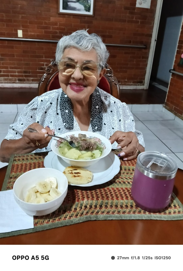 Una mujer sonríe sentada a una mesa, sosteniendo una cuchara sobre un plato de comida, con una taza y fruta cerca.