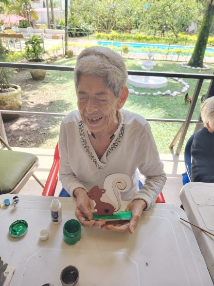 Una persona sonríe sentada a una mesa al aire libre, sosteniendo un dispositivo portátil verde con pequeños objetos cerca.