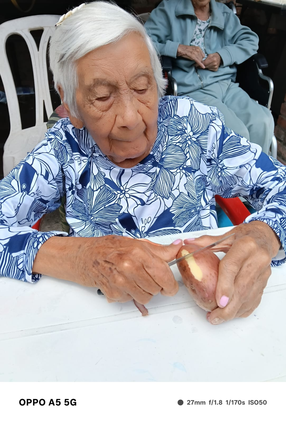 Una anciana con una blusa azul de flores corta una fruta en una mesa al aire libre.
