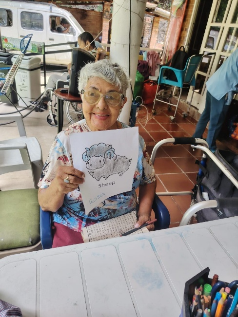 Una mujer sonriente sentada a una mesa sostiene un dibujo en una concurrida cafetería con terraza.
