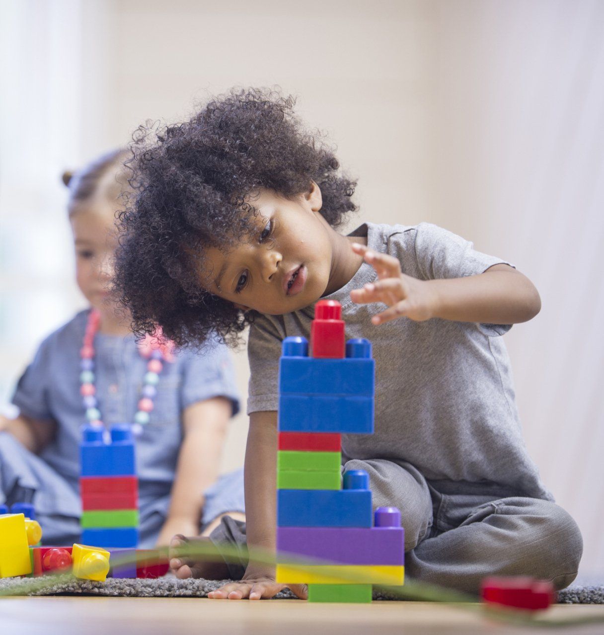 Children stacking colorful blocks on the floor, with one child concentrating on a tower in the foreground