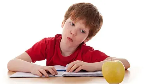 A young boy is sitting at a desk with a book and an apple.