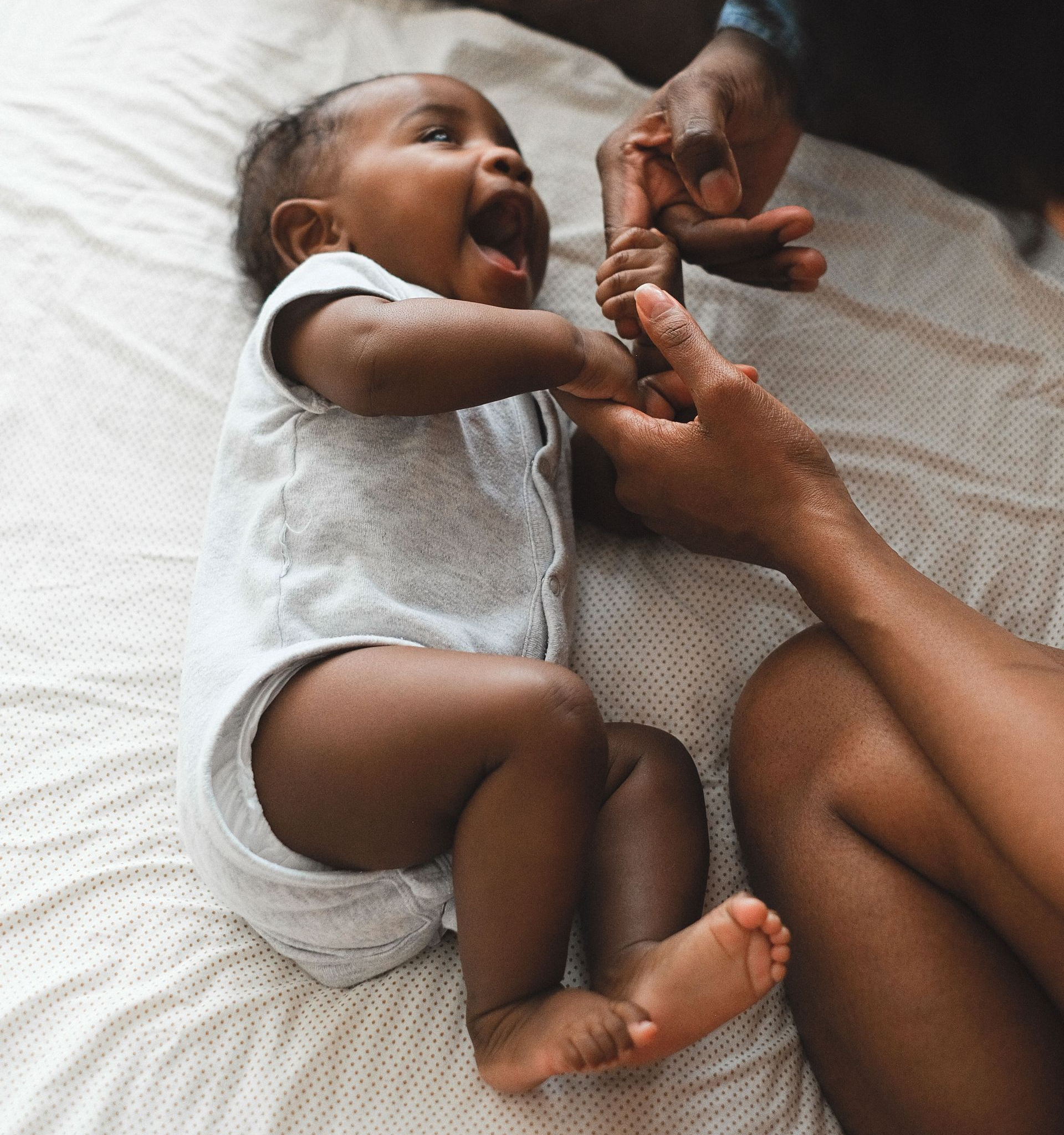 Baby lying on a bed, laughing while an adult gently holds their hands