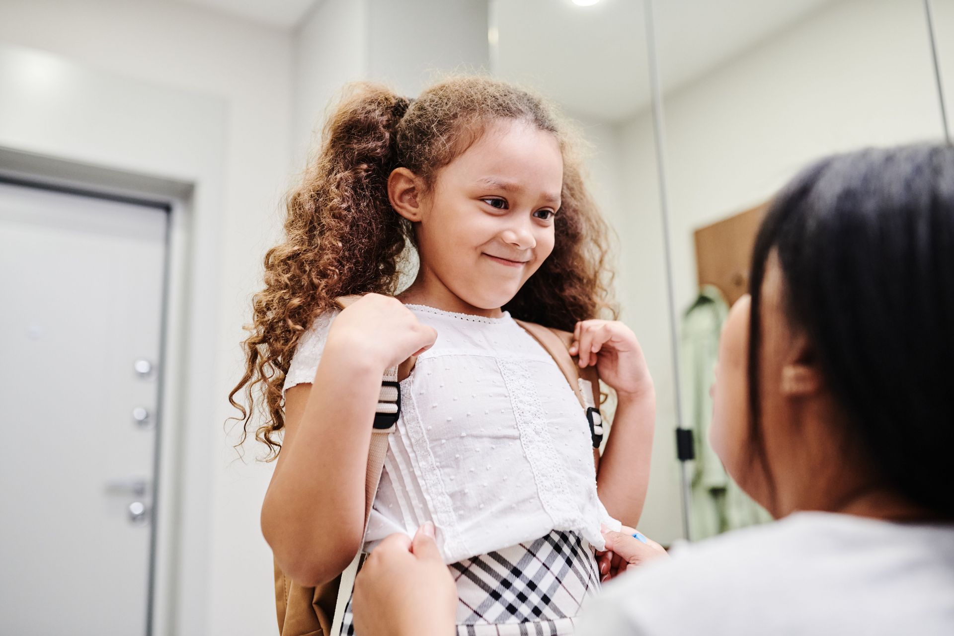 Child smiling while getting fitted with a backpack by an adult in a bright room