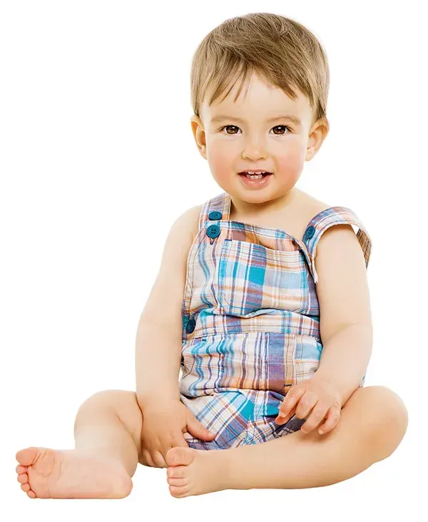A baby boy is sitting on the floor wearing plaid overalls.