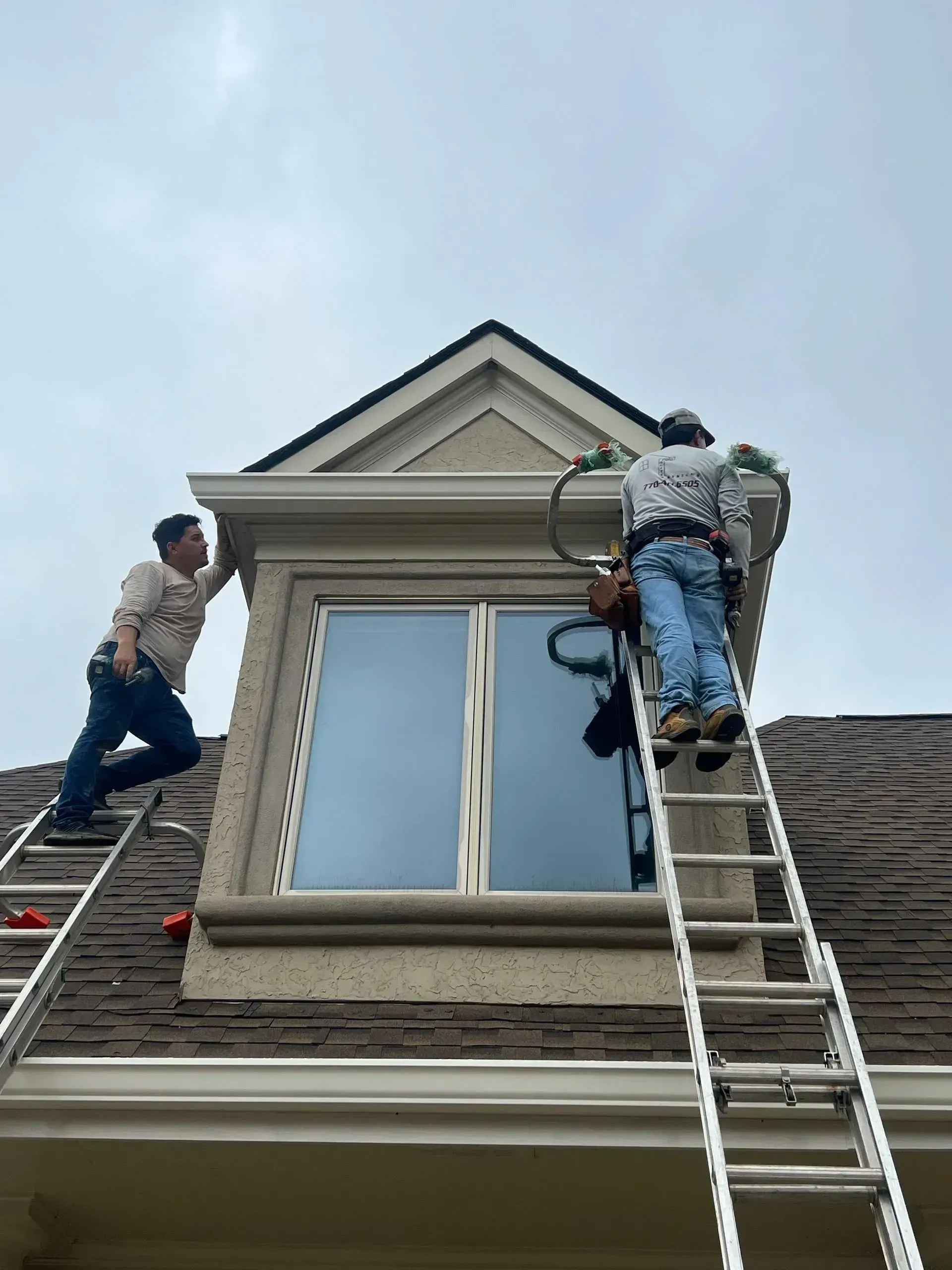 Two workers on ladders repairing the roof and siding above a second-story window.