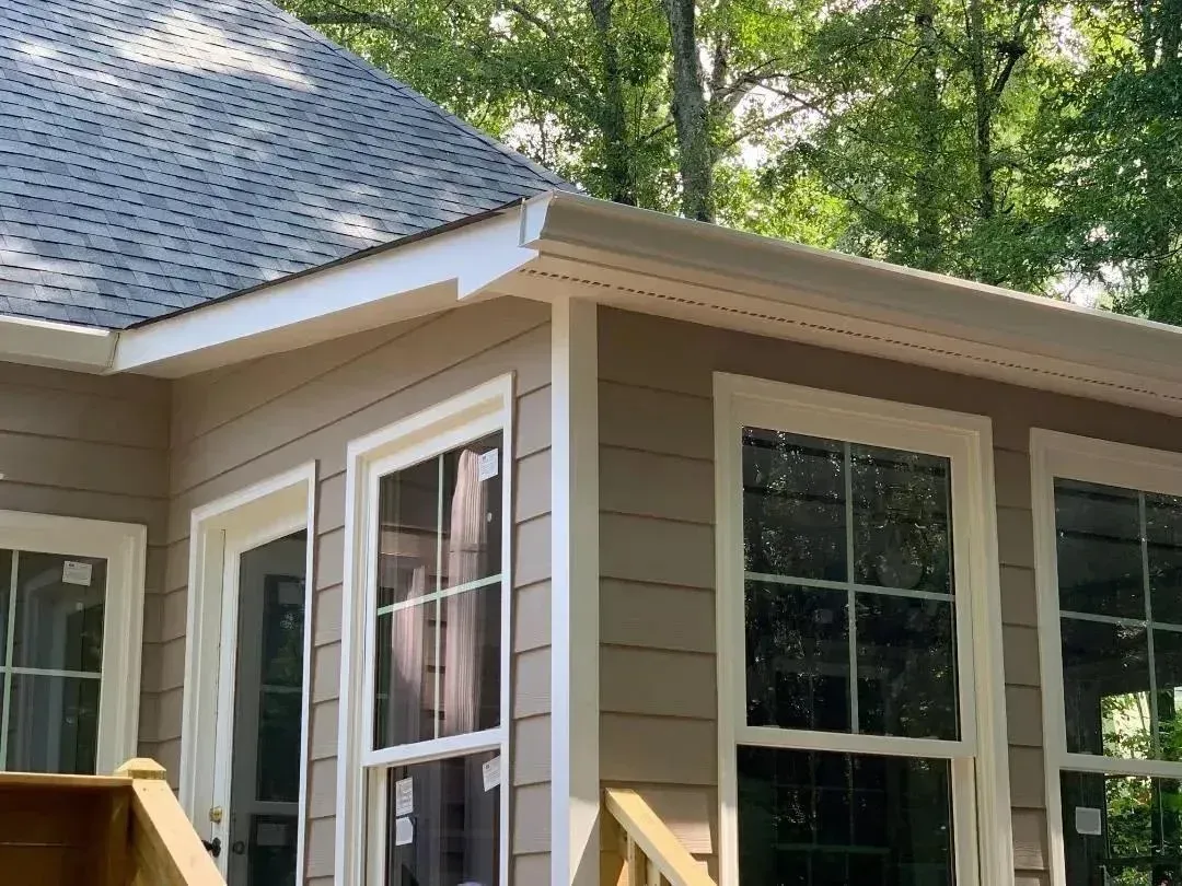 Tan house exterior with white trim, large windows, and a gray shingled roof surrounded by trees