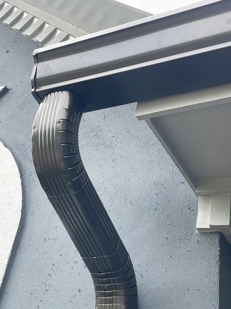 Black ribbed downspout under a roof gutter, running along a gray exterior wall.