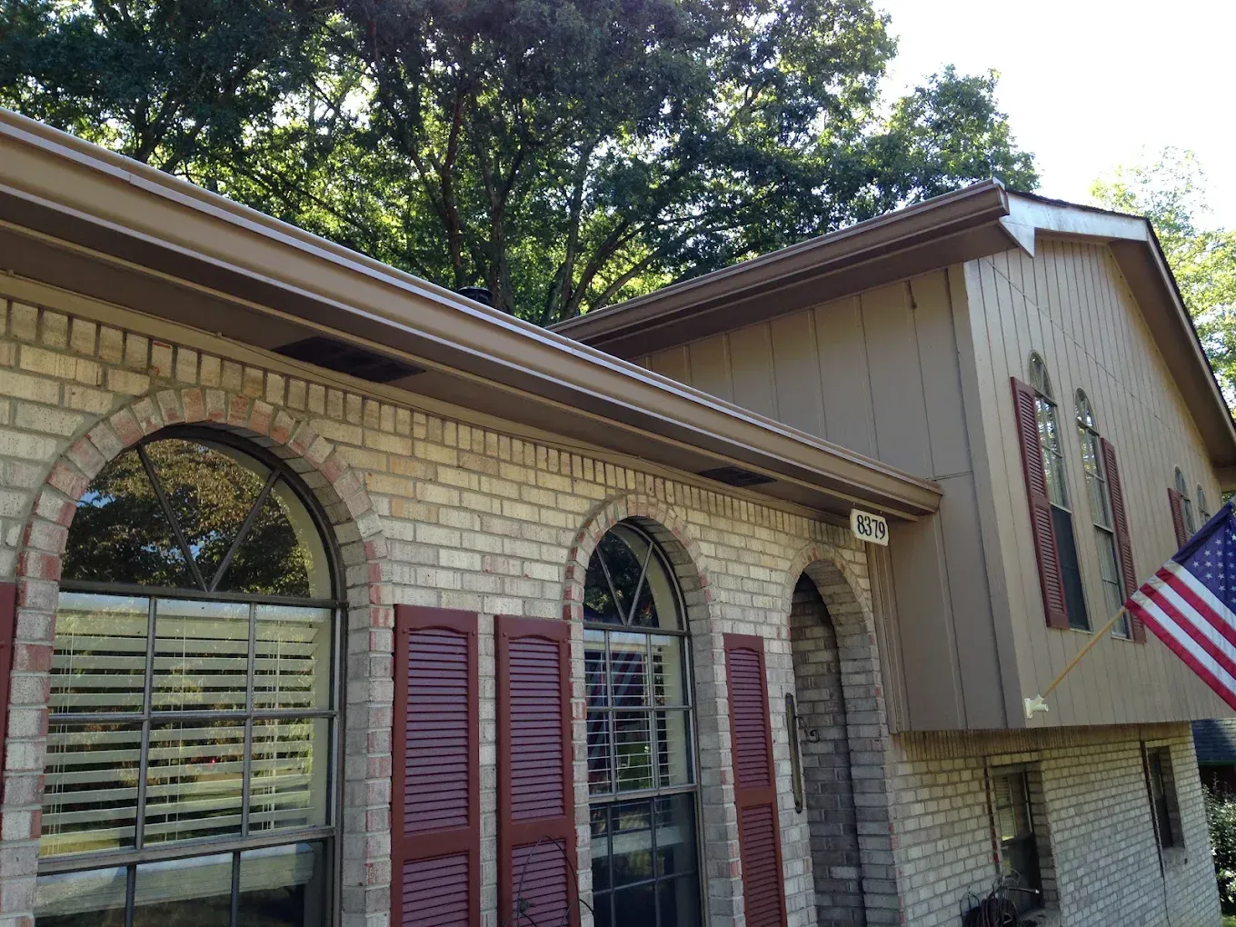 Suburban house exterior with brick facade, arched windows, shutters, trees, and an American flag