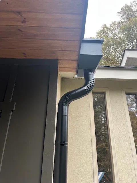 Black downspout and gutter on a house corner beneath a wooden roof overhang.