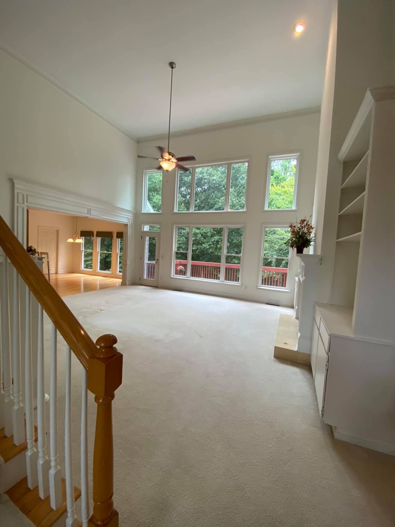 Bright open living room with vaulted ceiling, large windows, and beige carpet, viewed from a staircase.