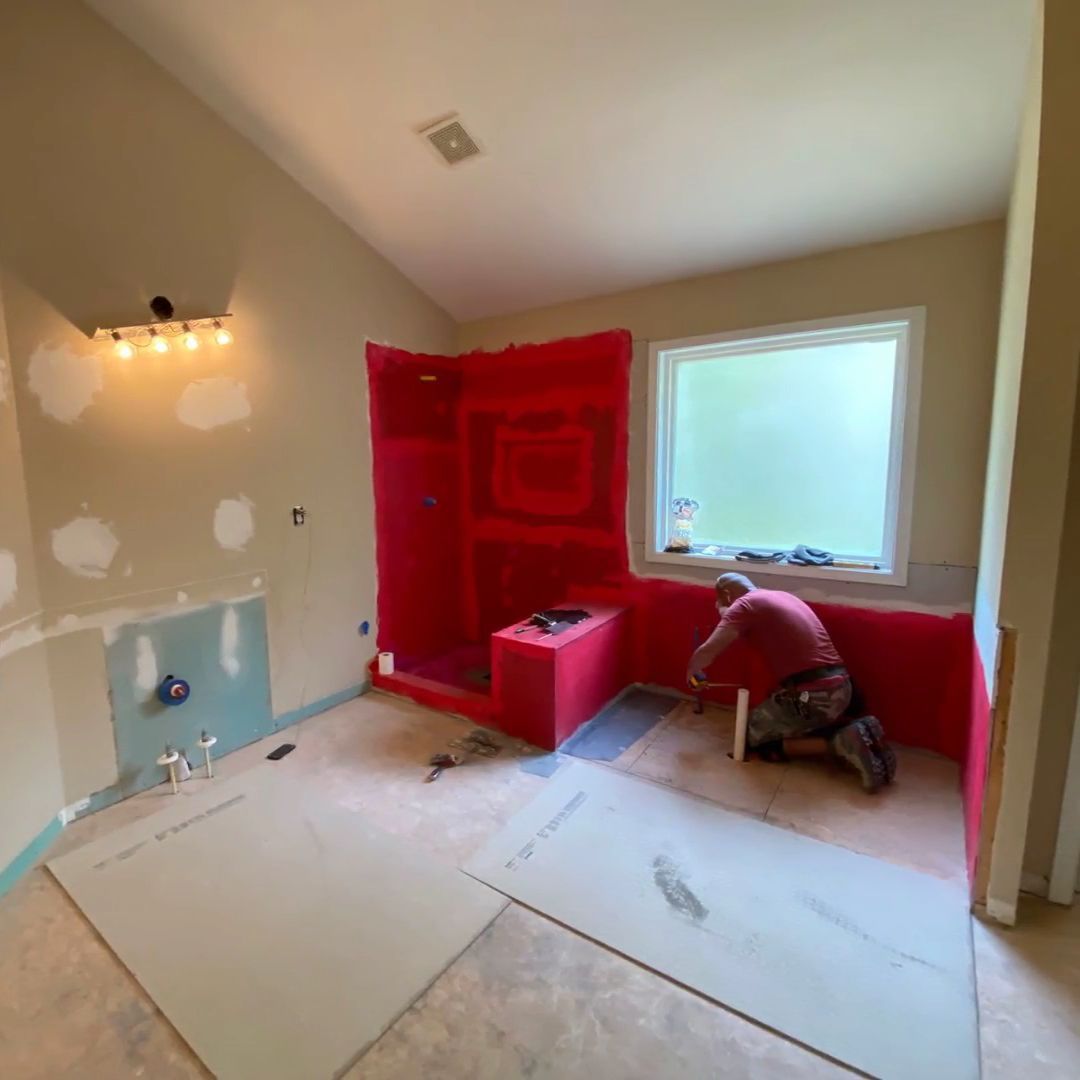 Person installing red waterproofing in a bathroom remodel near a window and bathtub alcove