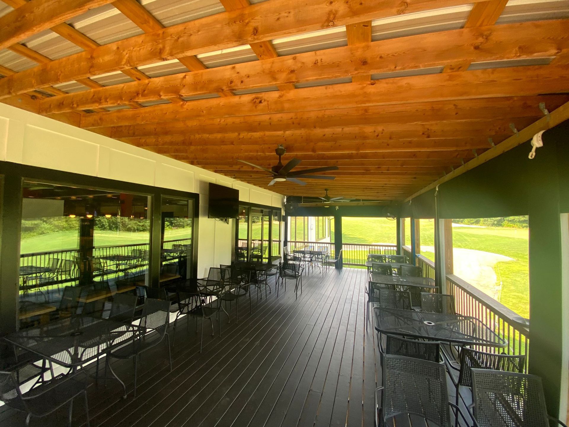 Covered outdoor patio with dark tables and chairs, wooden ceiling, and view of a green golf course.