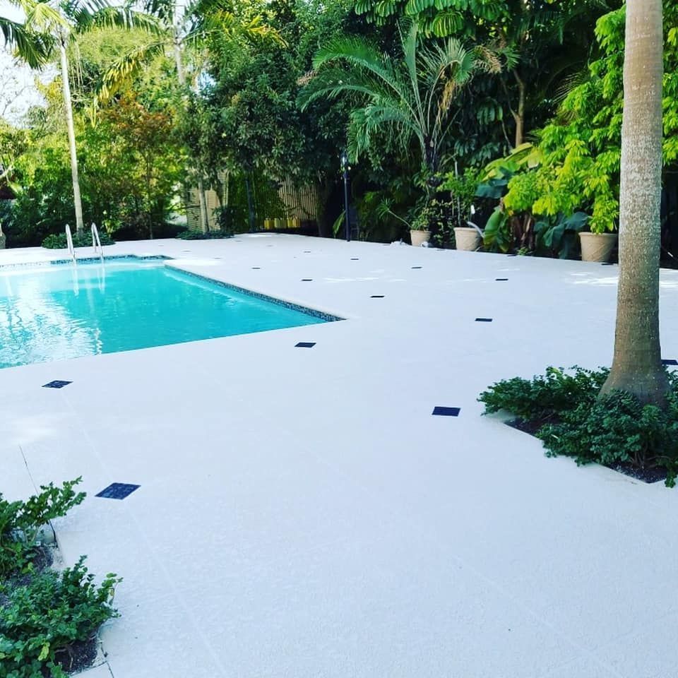 White pool deck beside a turquoise swimming pool, surrounded by lush tropical plants and palm trees